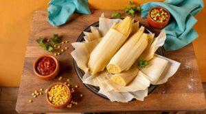 A rustic wooden table featuring a dish of tamales wrapped in corn husks, placed on a black plate. Beside the plate, small bowls contain bright yellow corn kernels, colorful mixed beans, and a rich red salsa. Fresh cilantro leaves and scattered corn kernels decorate the table, adding to the vibrant presentation. Blue cloth napkins add a pop of color against the warm, earthy background.