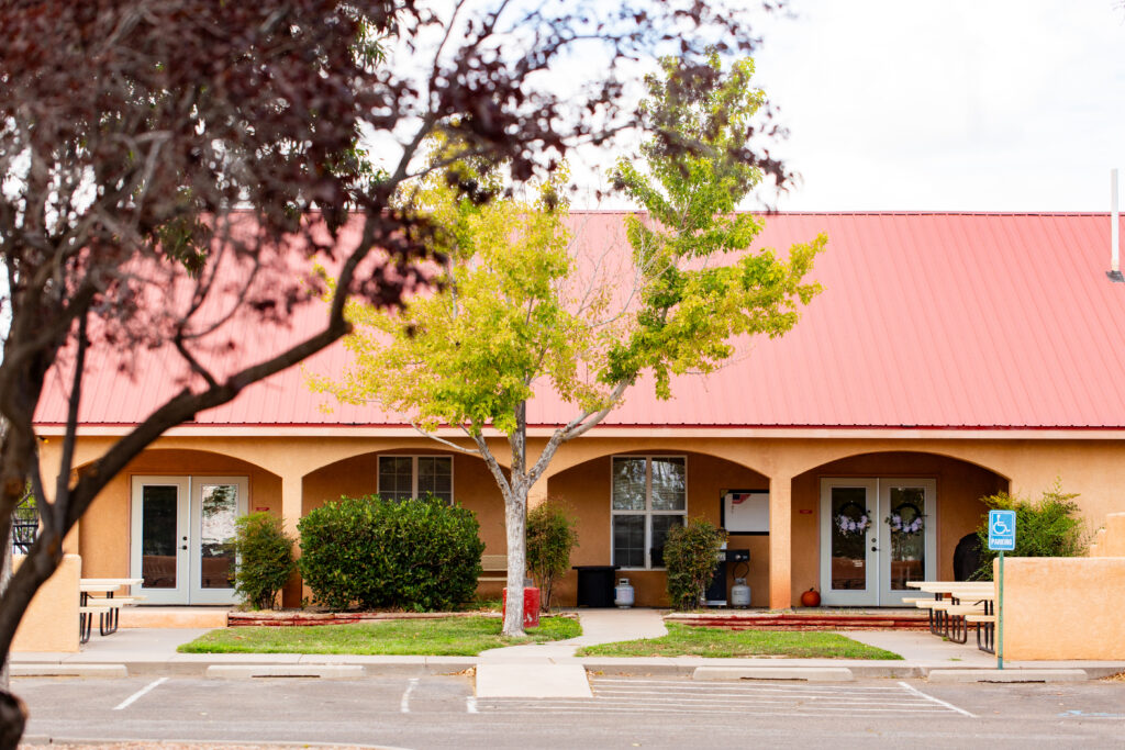 A building with a red metal roof and light brown stucco walls is partially obscured by trees with green and brown foliage in the foreground. The building has arched doorways with double glass doors, and windows flanking both sides. There are bushes and a small tree in front, and picnic tables on a concrete pathway leading to the entrance. A handicapped parking sign is visible in front, and there is a pumpkin near one of the doorways, suggesting a fall setting.