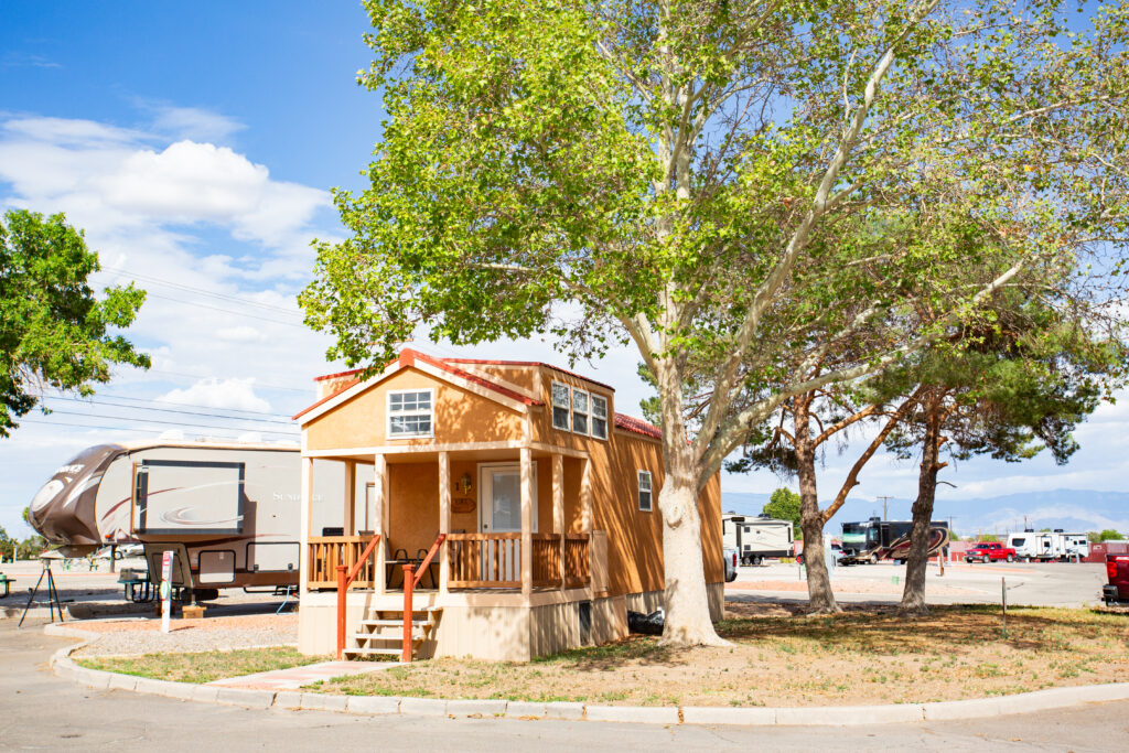 A small, beige tiny house with a red roof sits under the shade of a large tree. It has a small porch with two steps leading to the entrance. Several RVs and trailers are parked in the background in a spacious, sunlit campground. The sky is clear with a few fluffy clouds, and distant mountains are visible on the horizon, creating a serene and inviting outdoor setting.