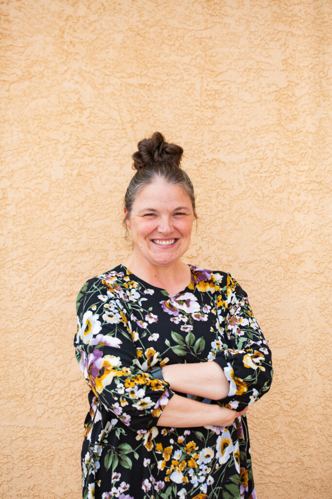A woman with her hair in a bun smiles confidently while standing with her arms crossed in front of a beige textured wall. She is wearing a black dress adorned with a vibrant floral pattern featuring yellow, white, and purple flowers. The warm tones of the wall complement the colors in her dress.