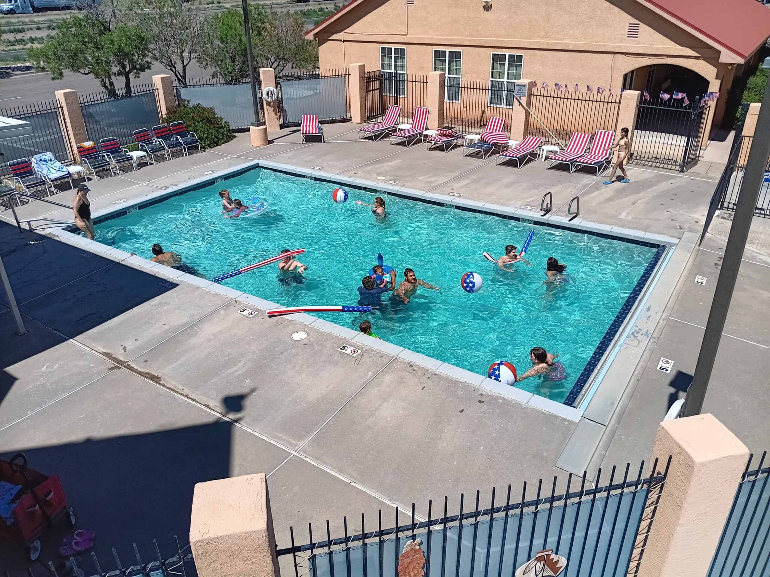A bird's-eye view of an outdoor swimming pool with about ten people enjoying a sunny day. Some adults and children are in the pool, playing with beach balls and pool noodles. The area surrounding the pool is paved, and there are red and white striped lounge chairs on the far side, some occupied by people sunbathing. A small building with a red roof is in the background. The pool is enclosed by a metal fence, with trees and an open field visible in the background. The scene conveys a relaxed, summertime atmosphere.