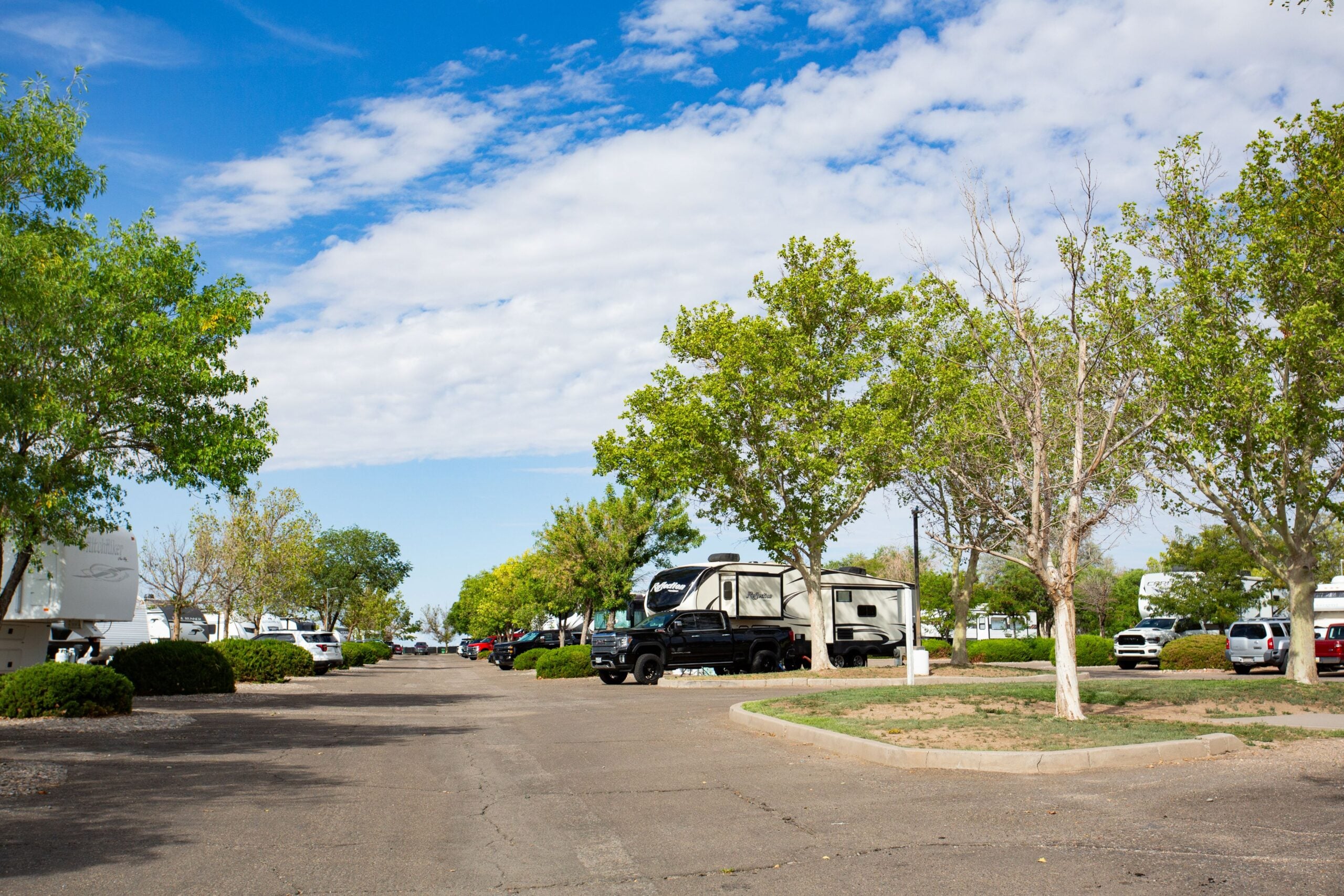 A sunny RV park with a clear blue sky and scattered clouds. The scene features a paved road lined with lush green trees and parked RVs on both sides. Some RVs have trucks parked next to them. The area is well-maintained with trimmed bushes and open patches of grass. The overall atmosphere is serene and inviting, ideal for outdoor leisure and camping.