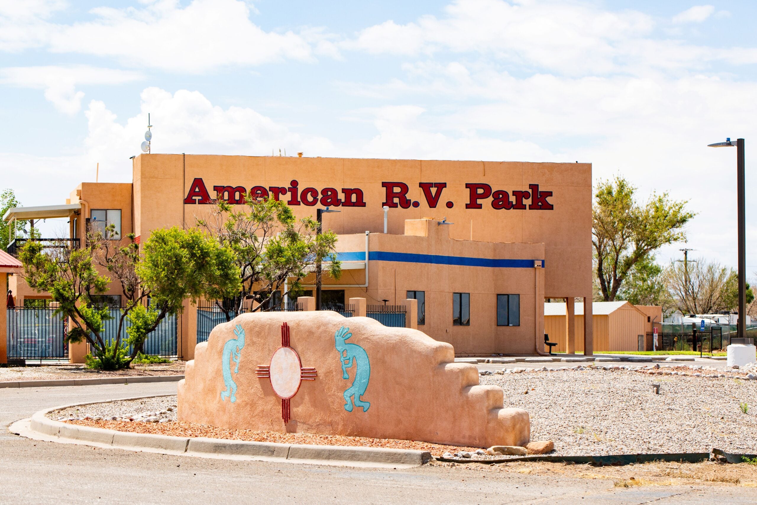 A beige building with "American R.V. Park" written in red letters; a decorative southwestern wall with turquoise kokopelli figures and the Zia sun symbol stands in the foreground.