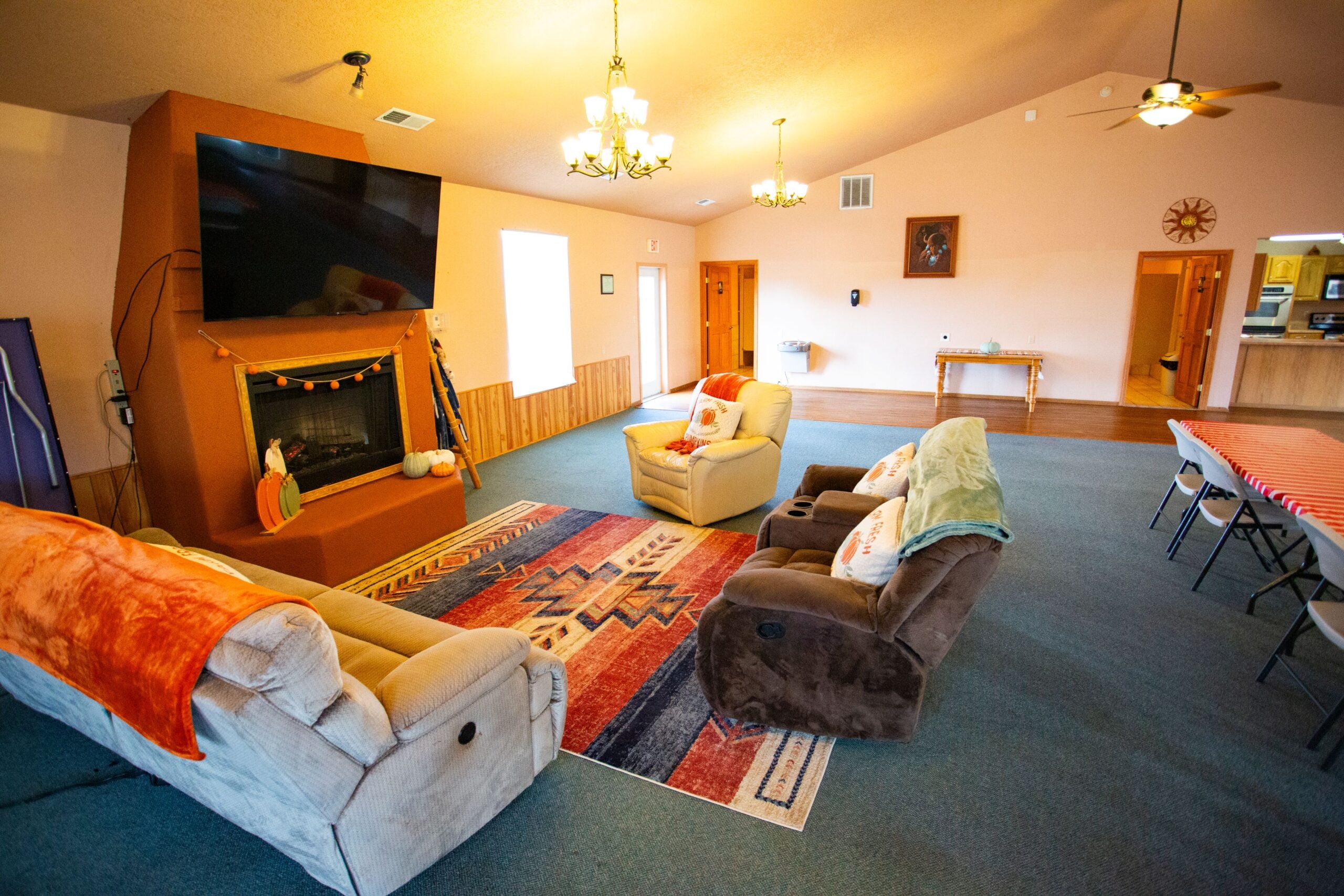 A cozy living room with a warm, inviting atmosphere. The room features a large flat-screen TV mounted above a fireplace, which is adorned with festive autumn decorations including pumpkins and an orange garland. In front of the fireplace, there are three recliner sofas covered with soft blankets and decorative pillows. A colorful area rug with geometric patterns adds vibrancy to the space. The walls are painted peach with wooden paneling on the lower half, while a ceiling fan and several light fixtures provide illumination. In the background, there are doors leading to other rooms and a table along the wall. On the right side, a long table is covered with a red and white striped tablecloth, accompanied by folding chairs.