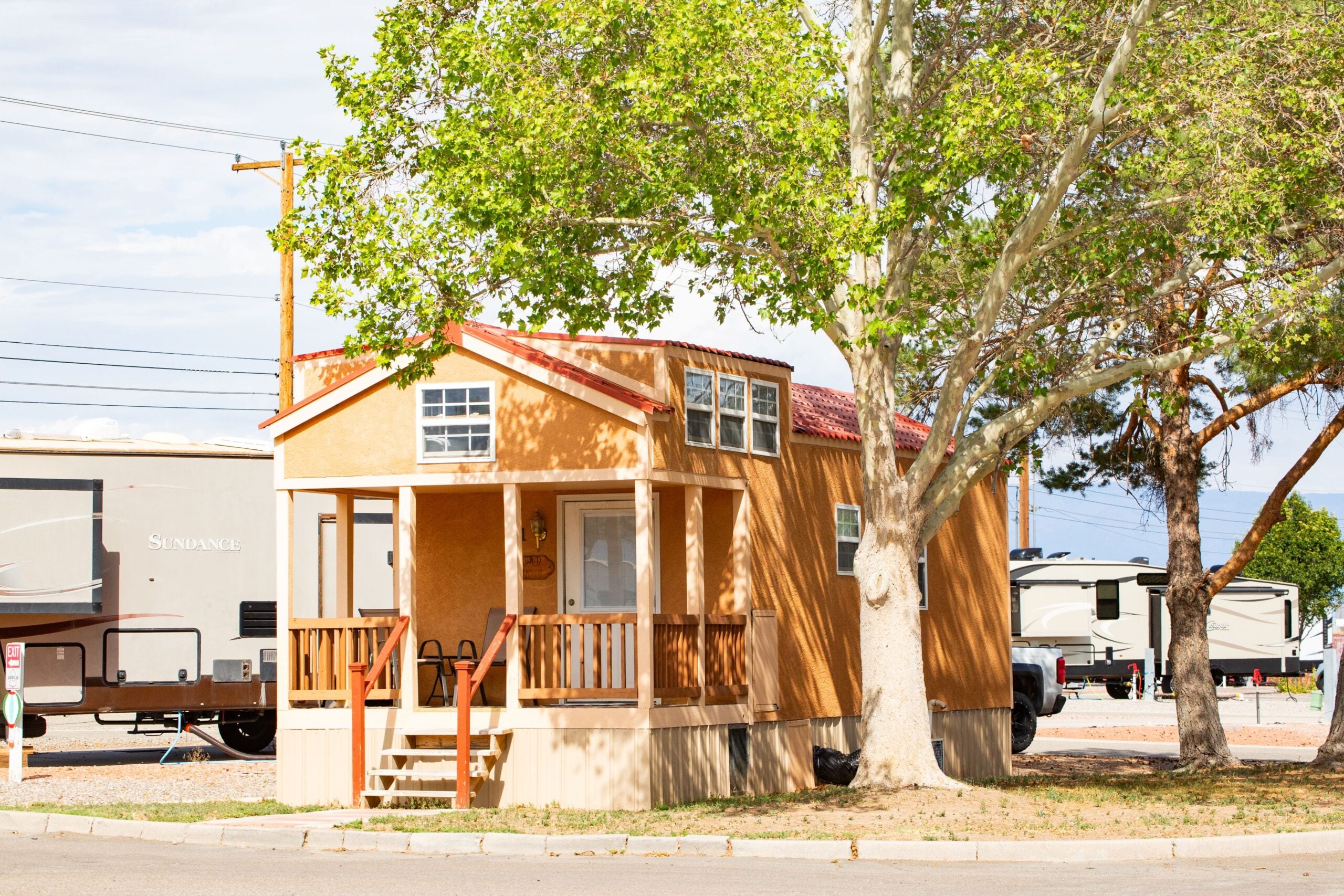A small tan tiny house with a front porch is parked near trees and surrounded by larger RVs in a lot.