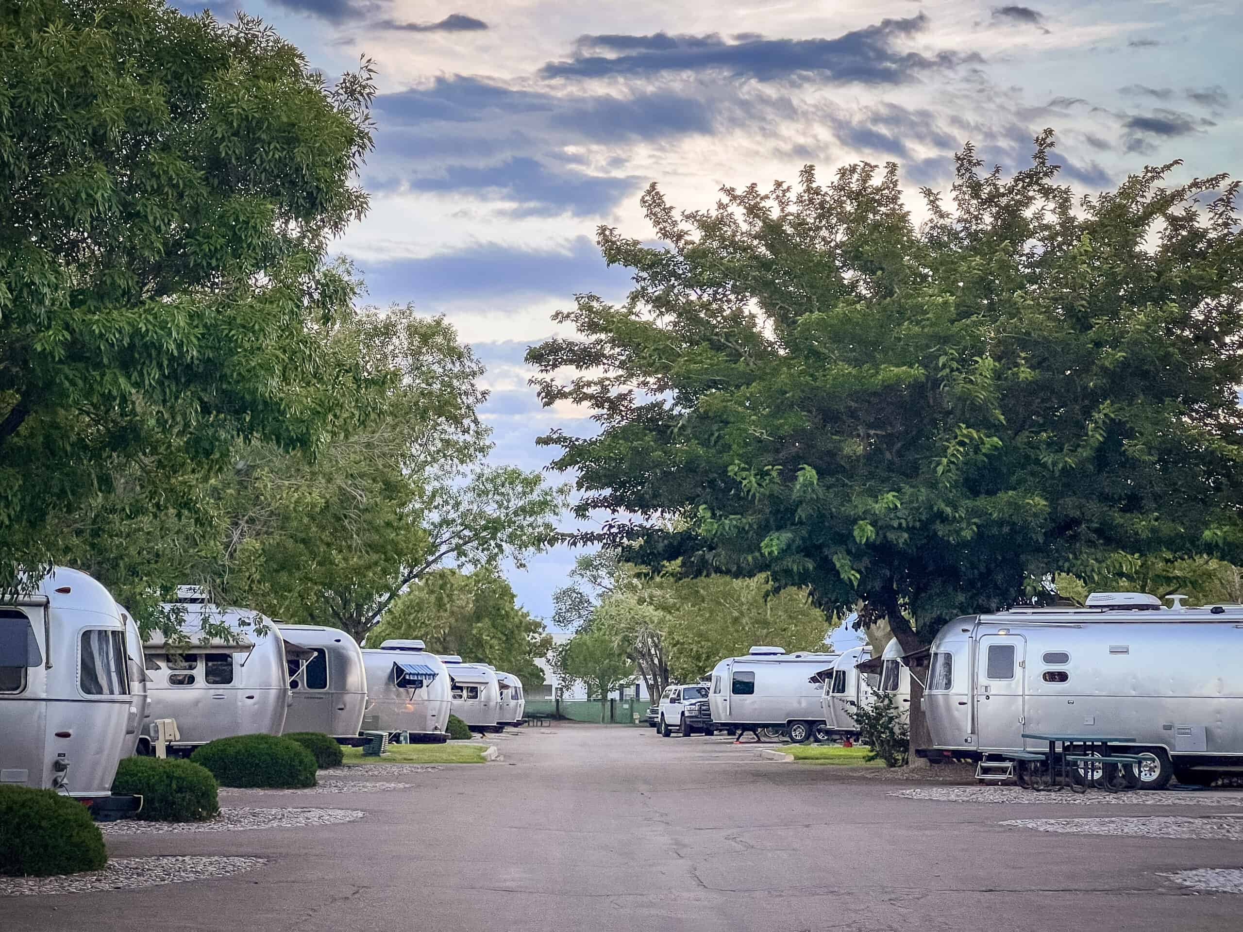 A scenic view of a well-maintained RV park with rows of shiny, silver Airstream trailers lined up on both sides of a paved road. The area is lush with large green trees providing ample shade. The sky above is partly cloudy, with patches of light breaking through, suggesting either early morning or late afternoon. The landscape is calm and inviting, ideal for a peaceful camping experience.