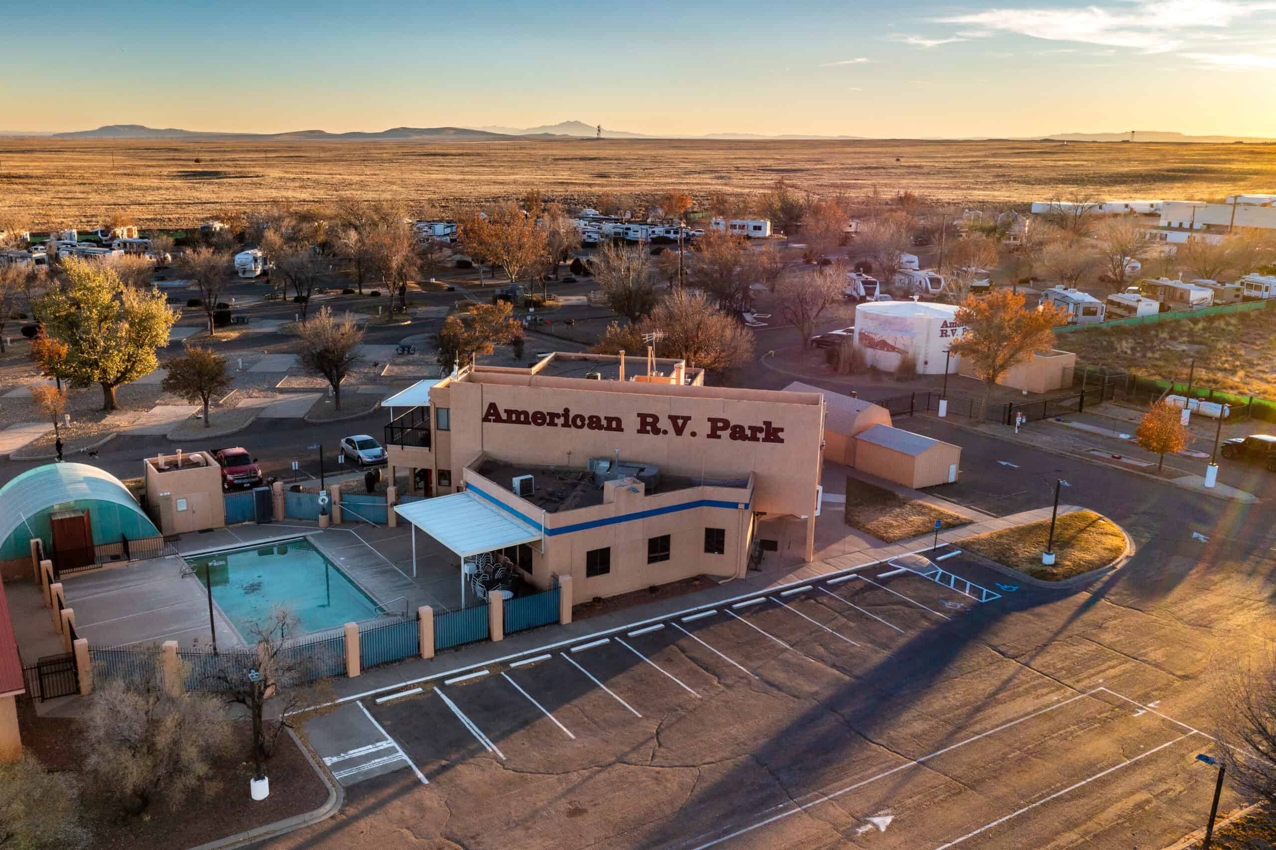 Aerial view of American R.V. Park with parked RVs, cars, swimming pool, and surrounding trees in a desert landscape at sunset.