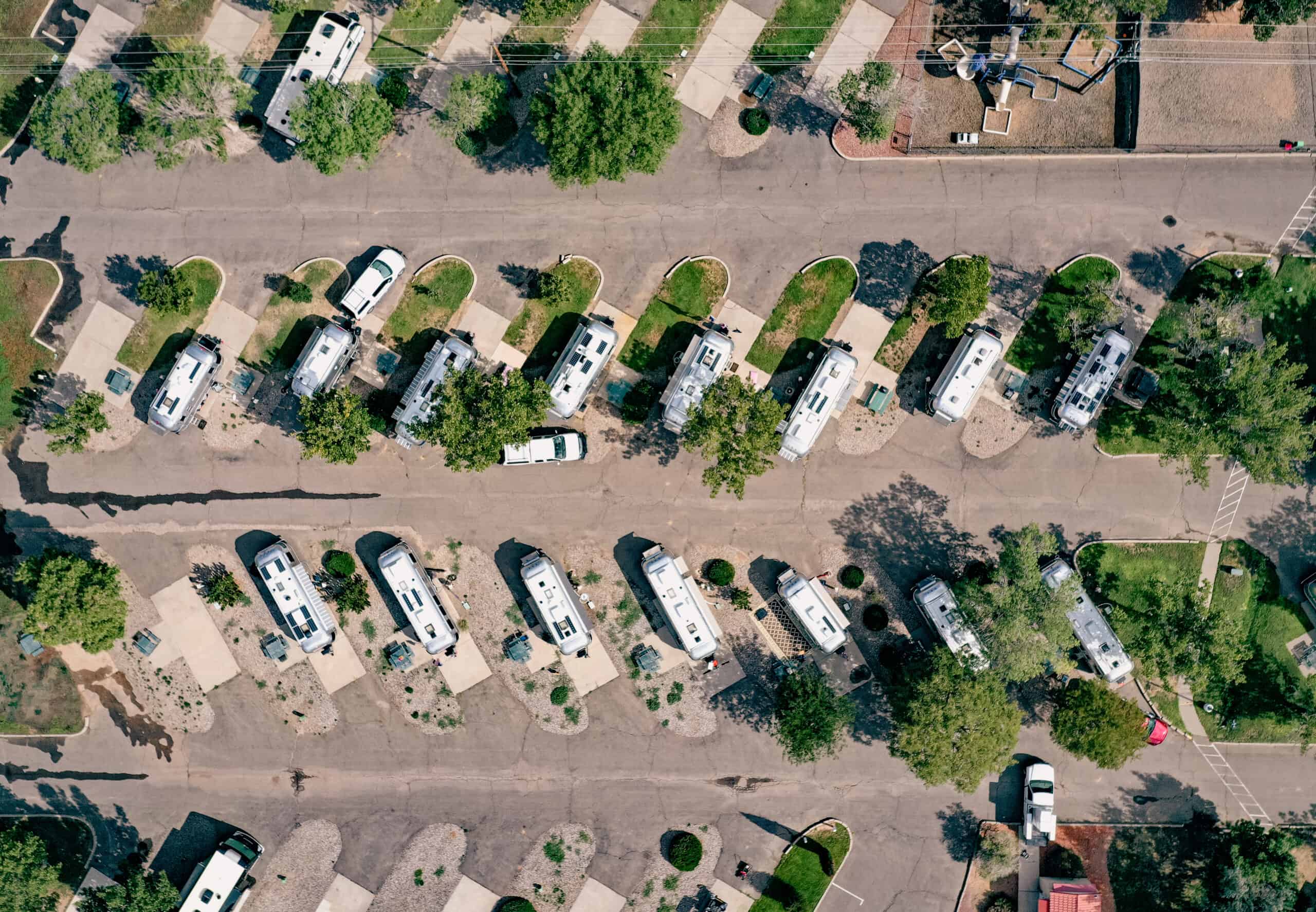 Aerial view of a well-organized RV park featuring multiple white recreational vehicles parked in designated spots. The parking spaces are arranged diagonally along paved roads and are separated by small, landscaped grassy areas with trees. Some accompanying vehicles are parked next to the RVs, and the overall scene is surrounded by more greenery and a few small structures, indicating a peaceful, structured environment.