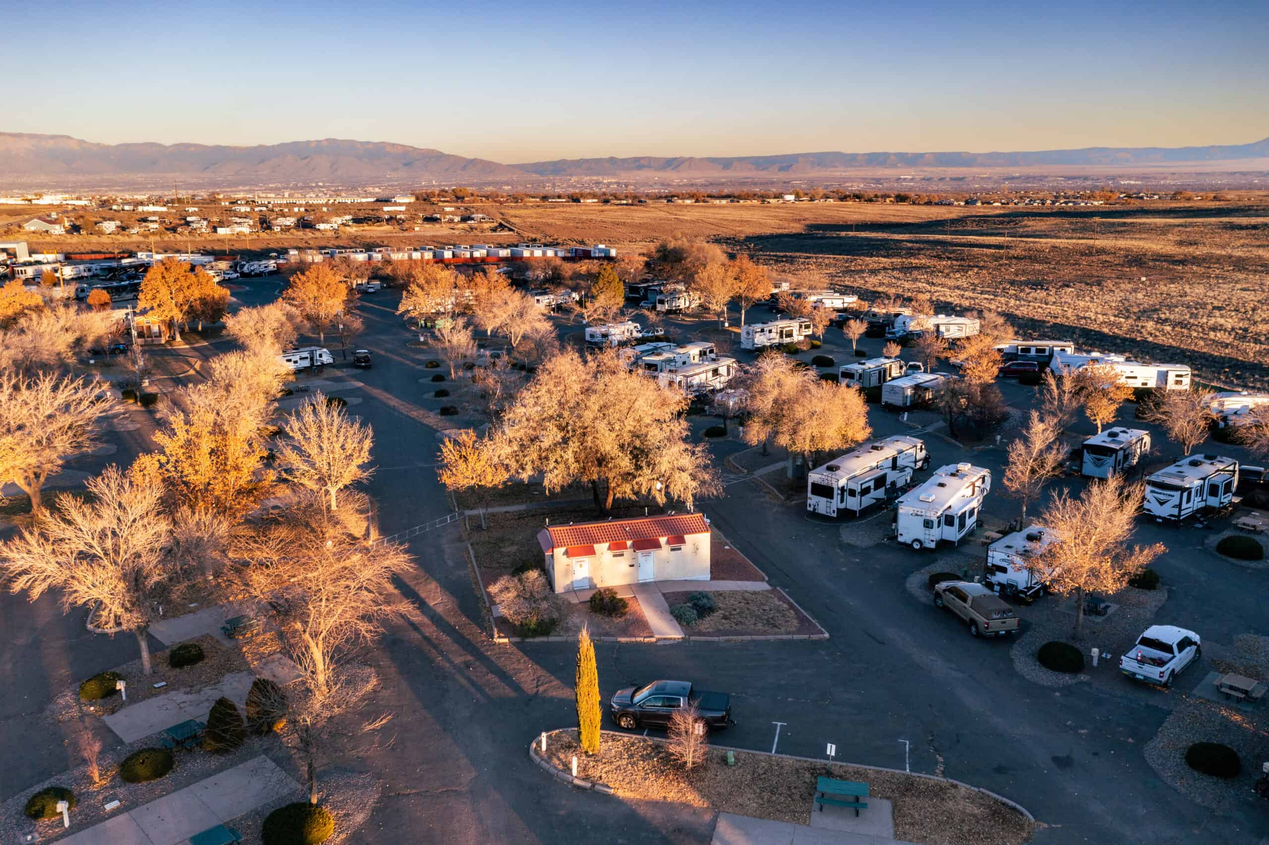 Aerial view of a spacious RV park set in a desert landscape during golden hour. Numerous white RVs are neatly parked along curving asphalt roads, which are lined with leafless deciduous trees casting long shadows. A small building with a red roof is centrally located amidst the RVs. Beyond the park, the expansive desert stretches towards distant mountain ranges. The clear blue sky is illuminated by the setting sun, enhancing the warm hues of the trees and landscape.