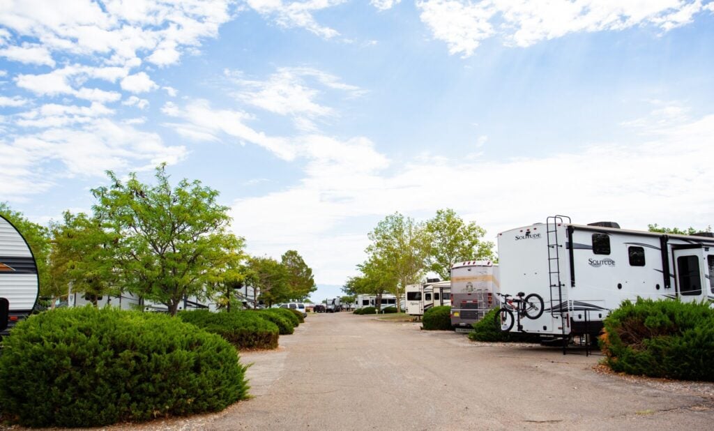 A paved road runs through an RV park with parked trailers, green bushes, and trees under a partly cloudy sky.