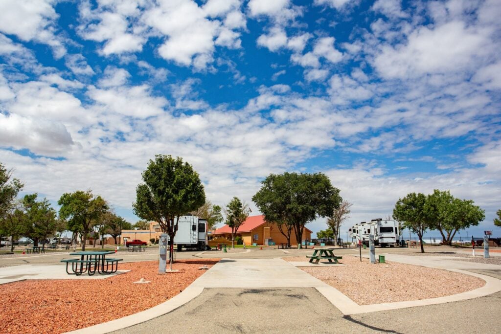 A paved RV park with several RVs, picnic tables, trees, and a building in the background under a partly cloudy sky.