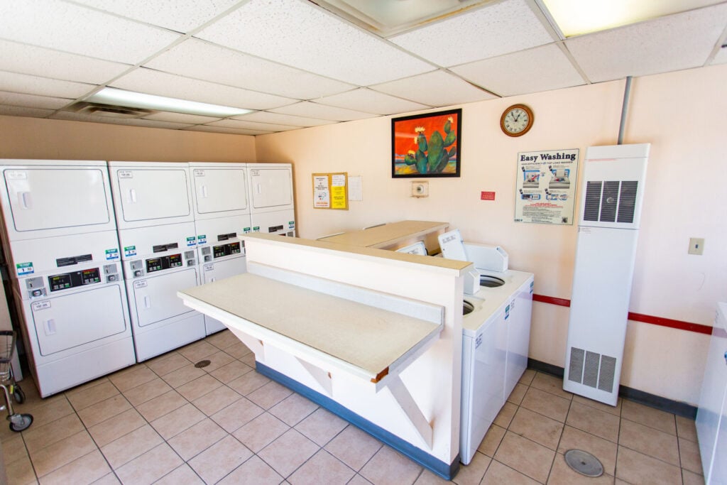 A laundromat with stacked dryers, top-loading washers, a folding counter, wall art, a clock, and instructional signs on cream-colored walls.