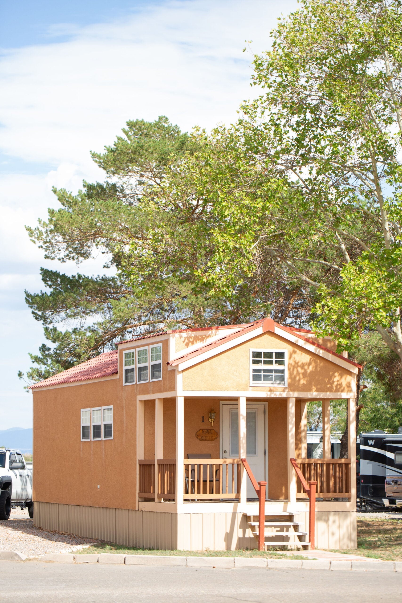 Small, tan tiny house with a red roof, front porch, white door, and multiple windows, situated under leafy trees on a sunny day.