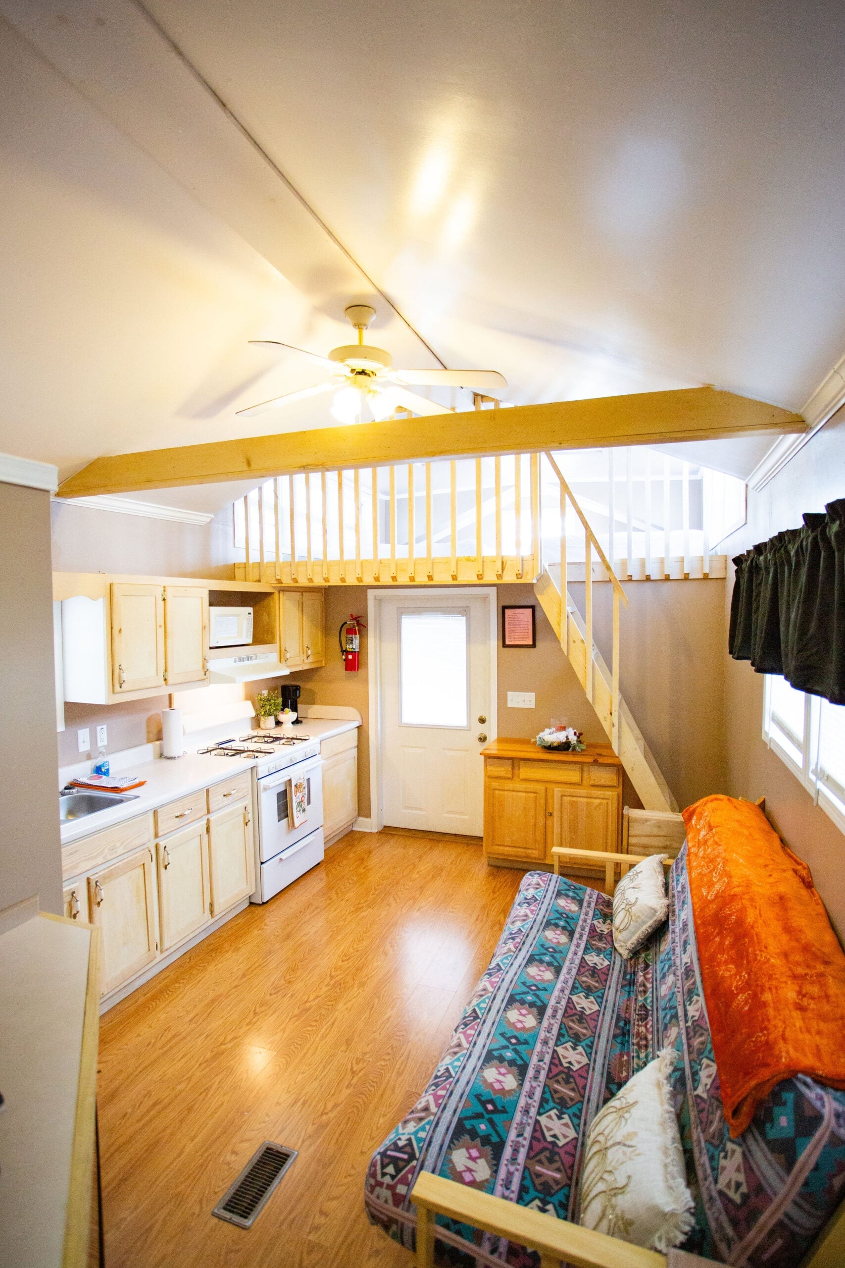 Small kitchen and living area with wooden floors, a patterned futon, white kitchen appliances, and a loft accessible by stairs. A ceiling fan and exposed beam are visible above.