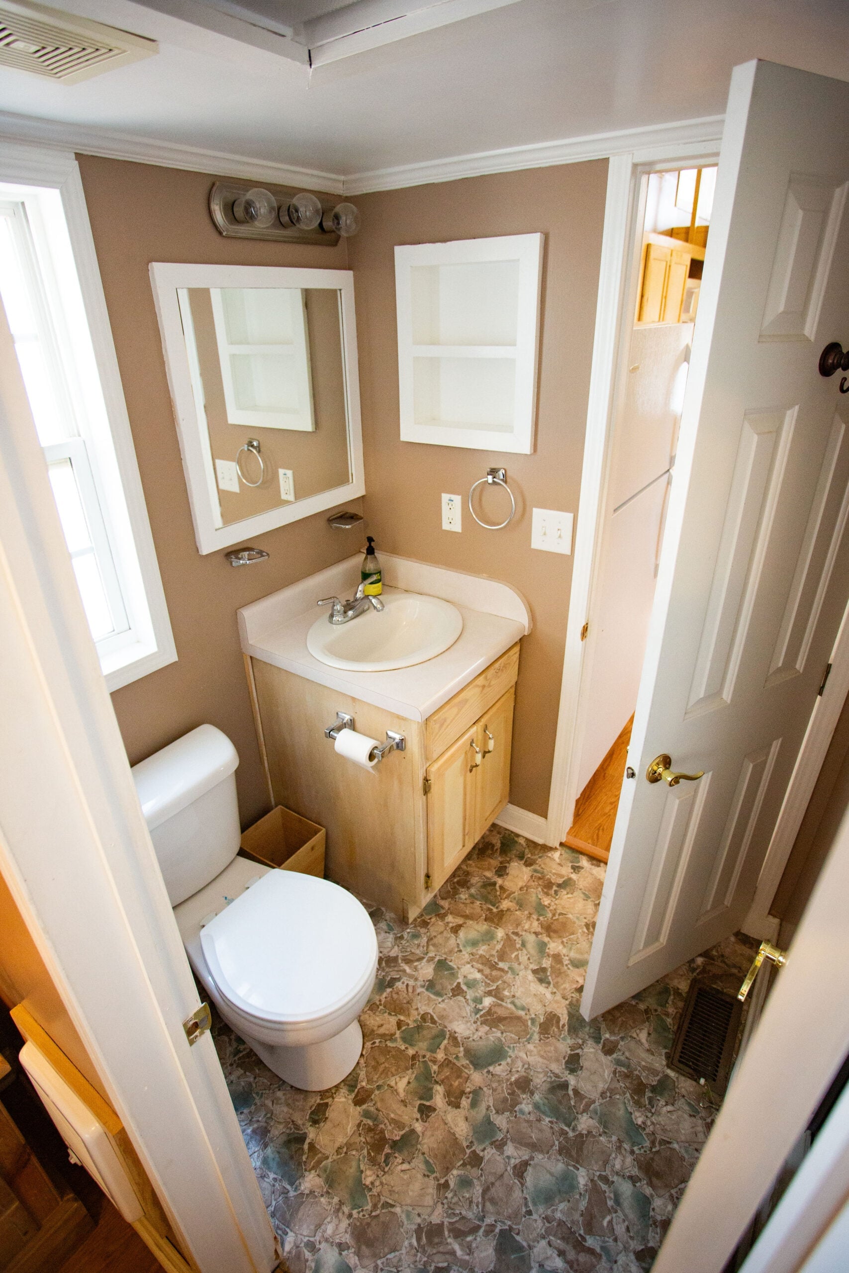 Small bathroom with tan walls, a white toilet, a sink with a light wood cabinet, a mirror, and a window. The door is open, showing part of an adjoining hallway with wooden stairs.