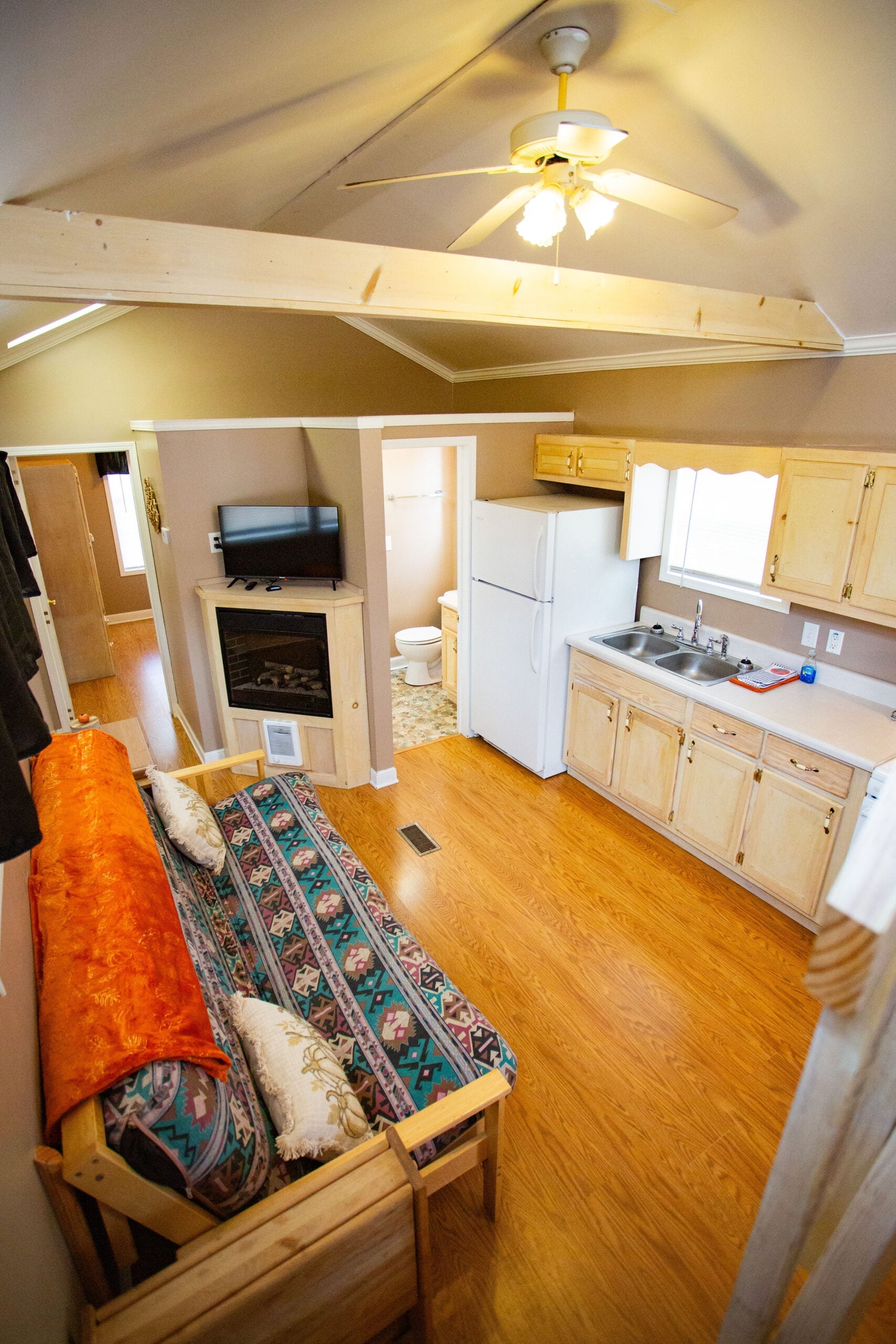 Small open-plan living area with a patterned futon, kitchenette, ceiling fan, fireplace, and view into a bathroom and other rooms; wood flooring and exposed ceiling beams.