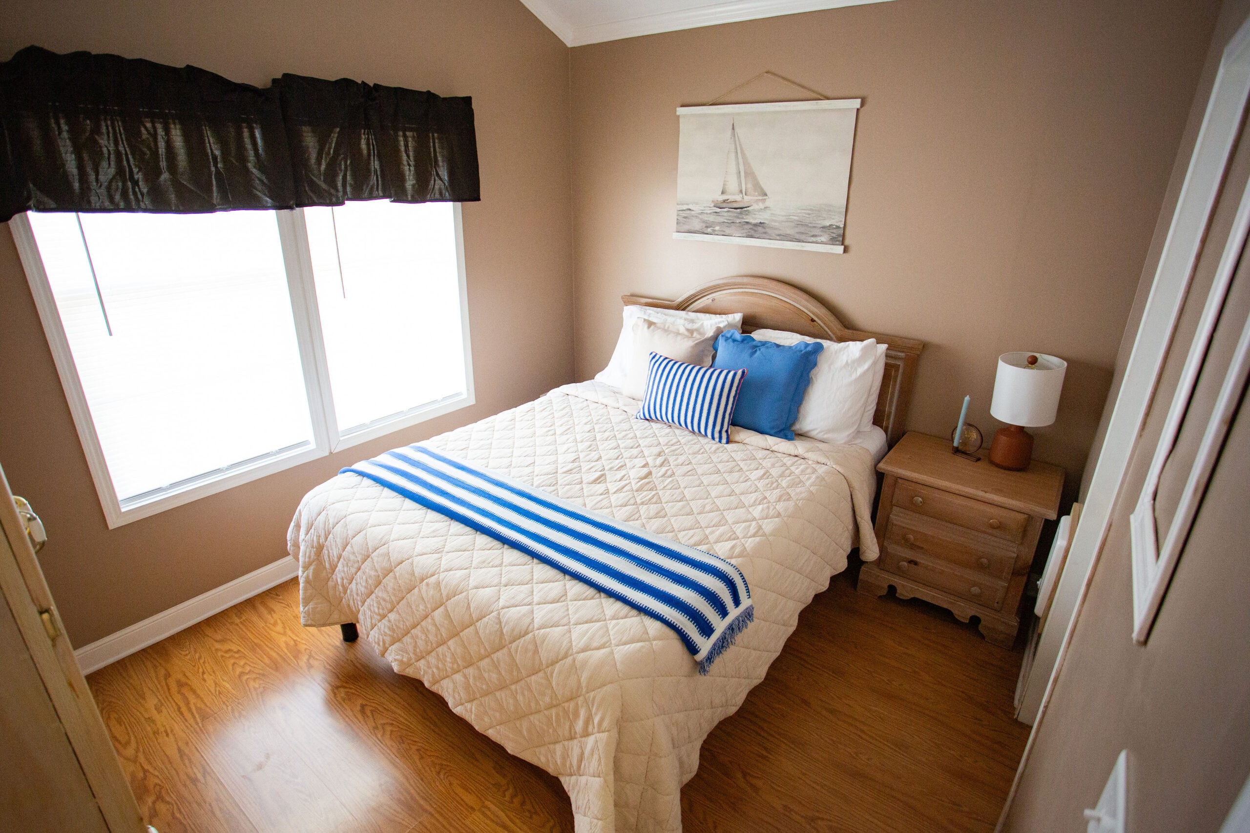 Small bedroom with a double bed, striped pillows and throw, wooden nightstand with lamp, window with brown curtains, and sailboat artwork on beige wall.