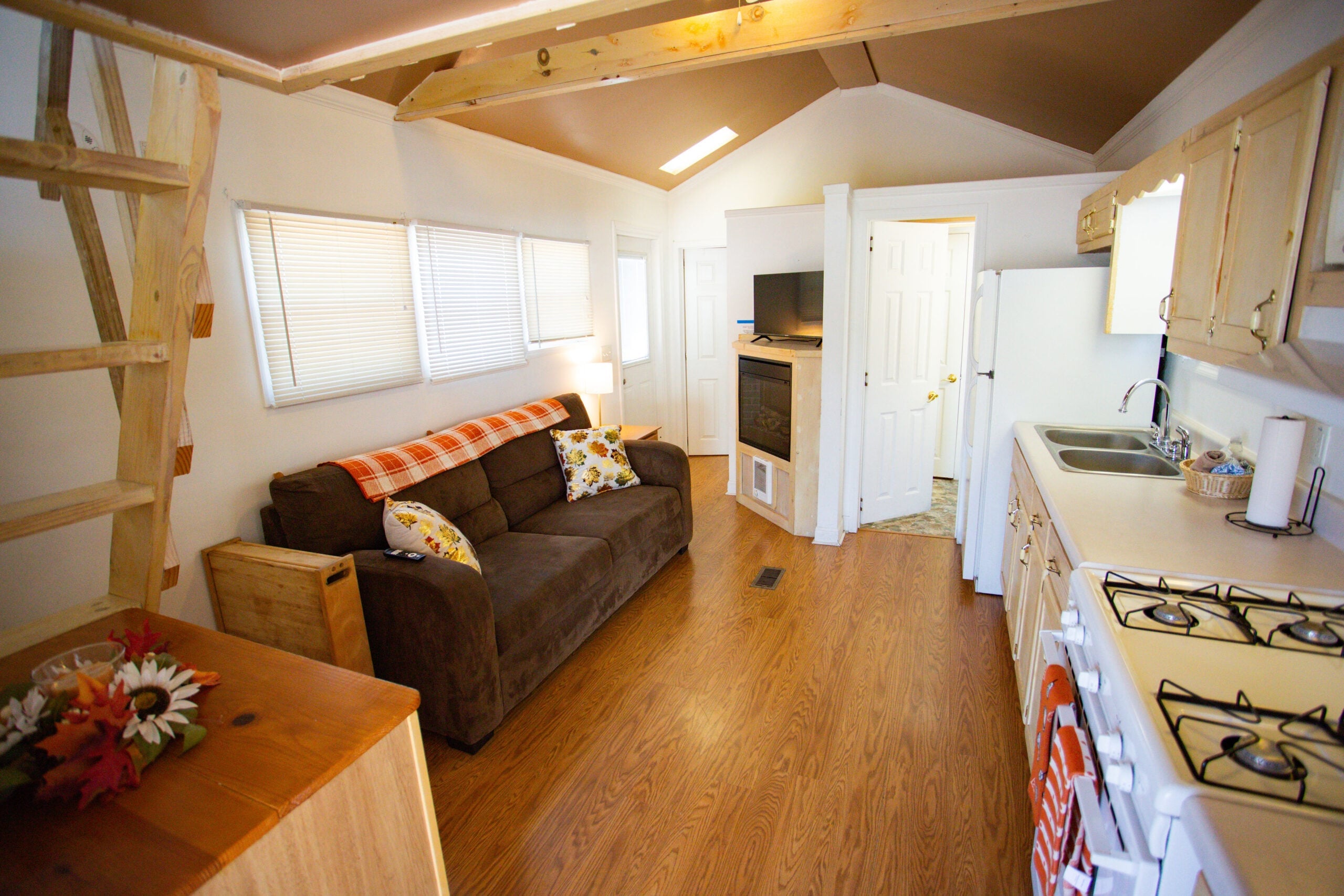 Small living area with brown couch, wooden floors, open kitchen, gas stove, TV, and exposed beams; stairs lead to loft above.