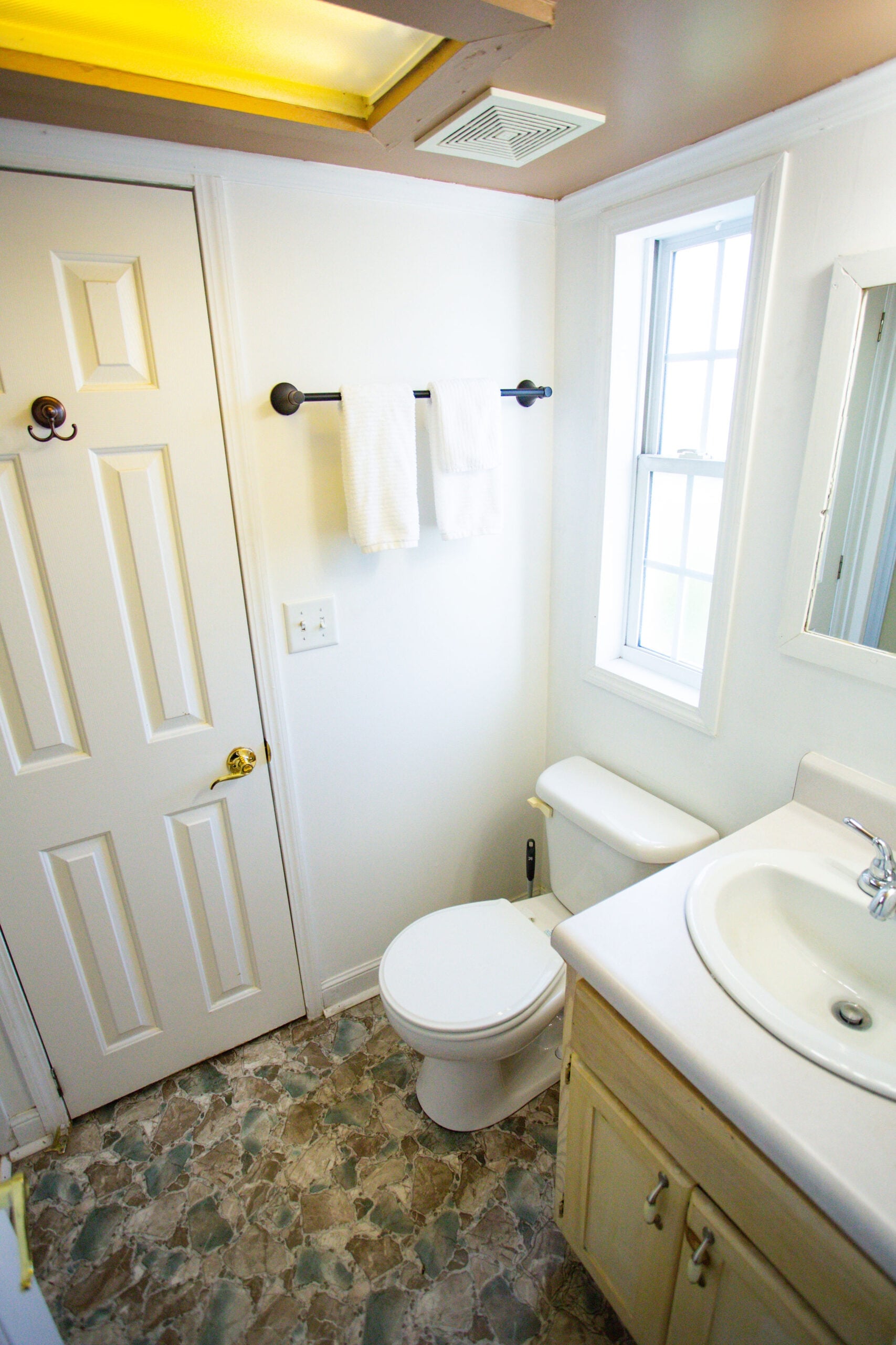 Small bathroom with a toilet, sink, white vanity, window, towel rack with two towels, and patterned tile flooring.