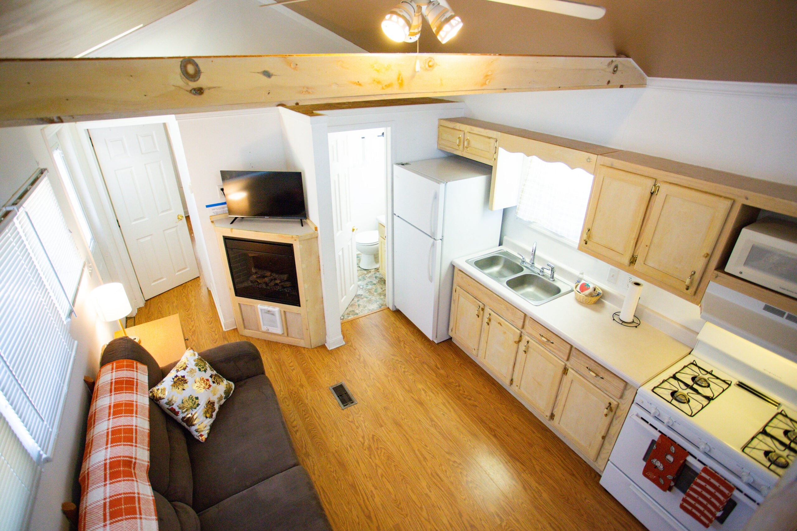 Small kitchen and living area with a sofa, refrigerator, stove, microwave, wall-mounted TV, and visible bathroom entrance; light wood flooring and cabinetry throughout.