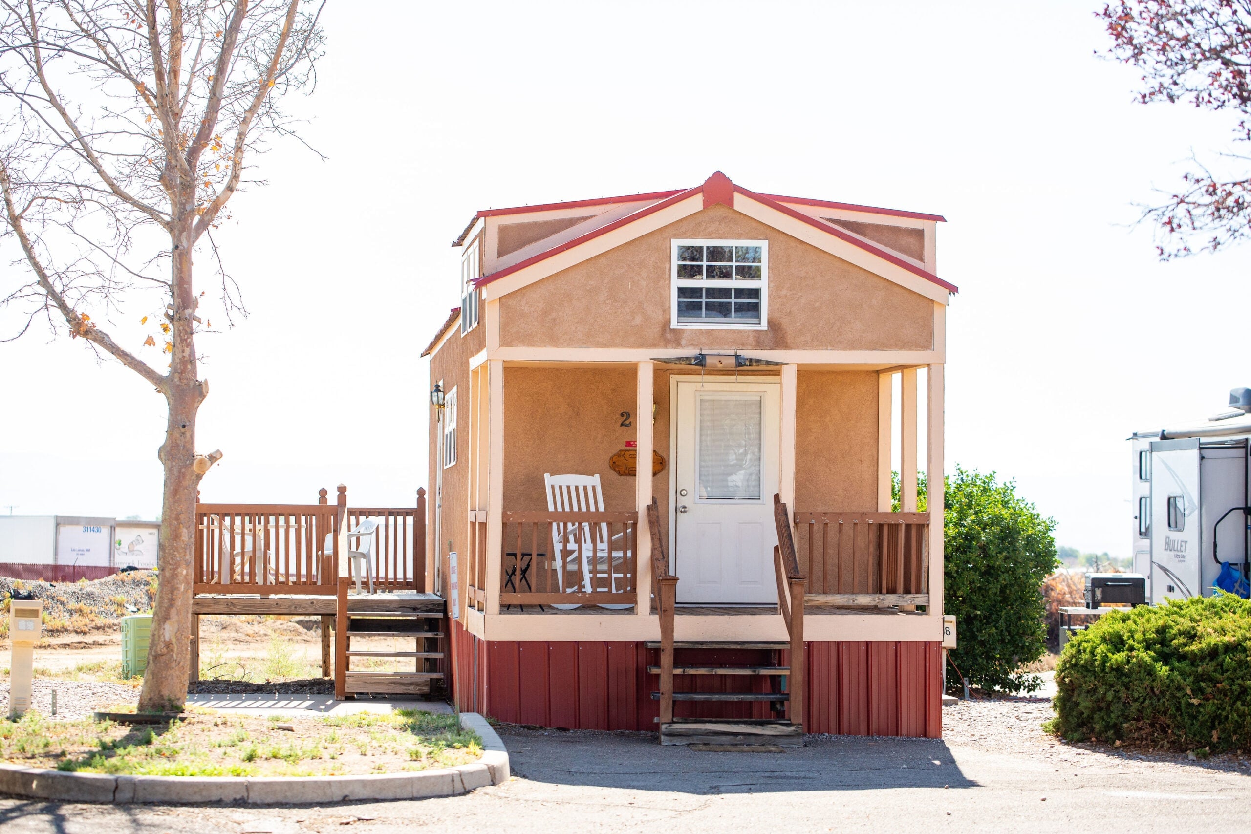 A small tan house with a front porch, rocking chairs, and steps leading to the door is shown. A leafless tree and a trailer are visible nearby under bright sunlight.