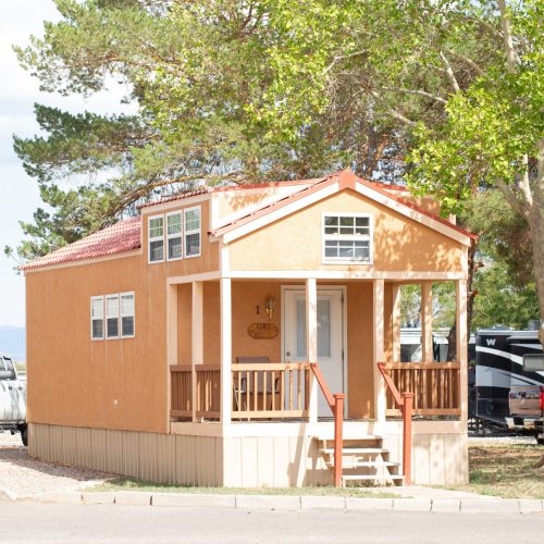 A small, cozy cabin with a warm, orange stucco exterior sits in a sunny, outdoor setting. The cabin features a covered porch with wooden railings and a set of steps leading up to the entrance. Several rectangular windows line the upper part of the cabin, letting in natural light. Surrounded by tall, leafy green trees, the cabin stands in a peaceful environment, with a few vehicles, including a white pickup truck and RVs, visible in the background. The setting has a campground-like feel, with clear skies overhead.