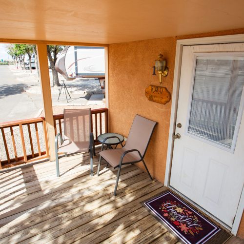 Small porch with two chairs and a glass table beside a white door with blinds. A doormat and a wall-mounted light fixture are also visible.