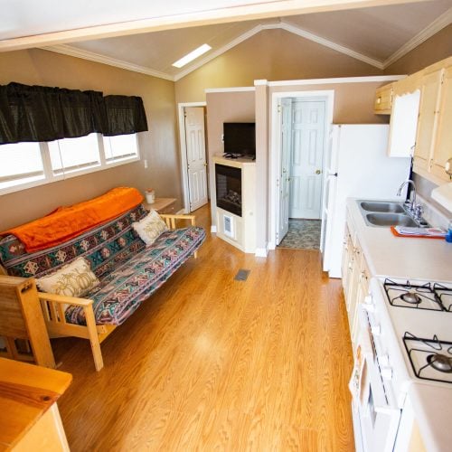 A cozy, compact living space with wooden flooring and neutral walls. On the left, there's a futon with a colorful patterned cover and an orange throw blanket, accompanied by decorative pillows. A narrow black curtain runs along the windows above the futon. In the foreground, an angled wooden table holds a small bouquet of flowers. To the right, a kitchen area features a white gas stove with four burners, a countertop with a double sink, and a microwave. A small fireplace with a TV on top is positioned against the far wall near a doorway, providing a glimpse into another room. The setting is warm and inviting with a blend of modern and rustic elements.