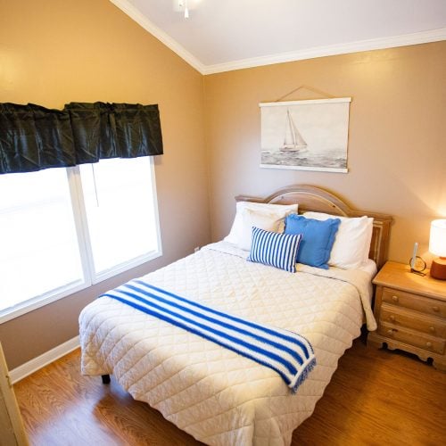 A cozy, small bedroom with tan walls and wood flooring features a neatly made bed with a white quilted bedspread. The bed is adorned with a blue and white striped pillow and matching throw blanket. A wooden headboard complements the matching wooden nightstand, which has a small lamp with a white shade and a clock. Above the bed hangs a nautical-themed painting of a sailboat. Two large windows with dark curtains provide natural light to the room.
