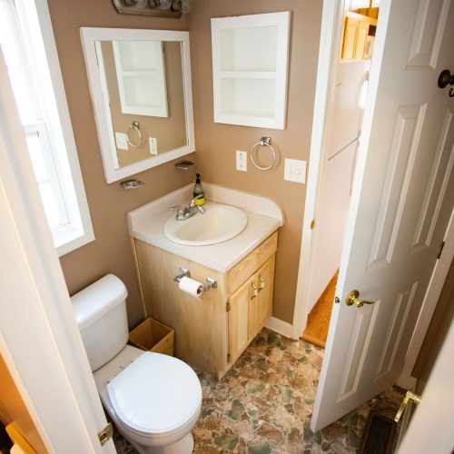 A small bathroom with beige walls and a patterned floor. The room features a white toilet and a wooden vanity with a white sink and silver faucet. Above the sink is a rectangular mirror with a white frame, and to the left, there's a cabinet with empty shelves. A round chrome towel ring is attached to the wall beside an outlet. Above the mirror, there is a light fixture with three bulbs. The bathroom door is slightly open, revealing an adjacent space with wooden stairs.