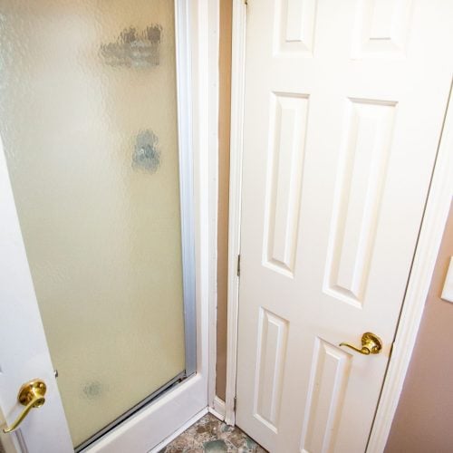 A small bathroom corner featuring a frosted glass shower door on the left with water spots visible. To the right, there is a white paneled interior door with a brass handle. Below, there's a floor with a mosaic tile pattern in shades of brown, gray, and green. The walls are painted in a warm beige color. The photo captures a compact space with a simple, functional design.