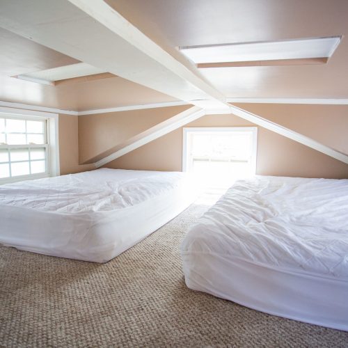 Attic bedroom with inclined ceilings painted in light brown, featuring two neatly made twin-sized beds with white bedding on a beige carpeted floor. Sunlight filters in through a low rectangular window at the far end, creating a cozy atmosphere. The room has minimal decor and two small windows along the side wall, contributing to the bright and airy ambiance.