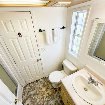 This image shows a small, well-lit bathroom with a white interior. The room features a white door with a dark metal hook, located on the left side. In the center, there is a toilet situated beneath a rectangular window that allows natural light to enter. On the right, a white countertop with a sink is visible, alongside a mounted rectangular mirror reflecting part of the room. A bottle of liquid soap is placed on the countertop. The room has a patterned floor with a mix of green and brown tiles. A black towel rack is mounted on the wall, holding two folded white towels.