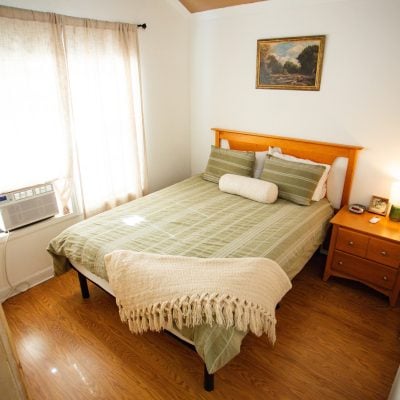 A cozy bedroom with wooden flooring features a neatly made bed against the wall. The bed has a light green plaid blanket, two matching pillows, and a white cylindrical cushion. A light beige, fringed throw is draped over the corner of the bed. To the right, a wooden nightstand holds a lamp with a soft green base, a picture frame, and a few small items. A painting hangs above the bed. Natural light filters through sheer beige curtains that cover a window with a visible air conditioning unit.