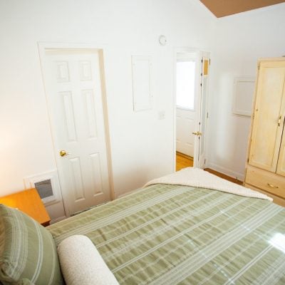 A cozy bedroom featuring a neatly made bed with green striped bedding and a white blanket folded at the foot. To the left, a wooden nightstand holds a small lamp, casting soft light on a nearby wall. Opposite the bed is a light-colored wooden wardrobe with two doors and brass handles. The room includes a white door on the left wall and another partially open door on the right wall leading to a hallway with wooden flooring. The walls are plain white, contributing to a clean and minimalist aesthetic.