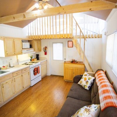 A cozy, small interior living space featuring an open-plan kitchen and living room area. The kitchen has light wood cabinets, a double sink, and white appliances, including a stove and microwave. The countertop is adorned with a basket of fruit, a paper towel holder, and a coffee maker. Adjacent to the kitchen is a dark brown sofa with colorful floral and plaid cushions, positioned beneath a window with white blinds. The room has warm wooden floors and is brightly lit. A staircase with wooden railing leads up to a loft area, visible above. The ceiling is angled, and a ceiling fan with lights is mounted above. A door with a window panel and a fire extinguisher are seen near the staircase.