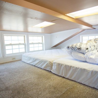 A cozy loft space featuring two mattresses side by side on a beige carpeted floor. The low ceiling is painted light brown with recessed lighting and two rectangular skylights, allowing natural light to illuminate the room. A row of four white-framed square windows lines the side wall, providing additional light. A plastic-wrapped item, possibly a bedding set, is placed on the mattresses. The overall atmosphere is airy and simple, with soft natural lighting.