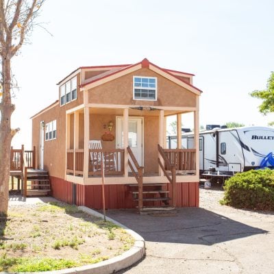 A small, brown tiny house with a gabled roof is situated in a lot surrounded by other RVs. The house has a front porch with a rocking chair, a white door, and a rectangular window above it. The porch is accessible by a wooden flight of steps. To the left of the tiny house, there is a bare tree and a patch of grass. To the right, a green bush and another tree can be seen, along with two large, white RVs. The scene is set in a sunny environment with a clear sky.